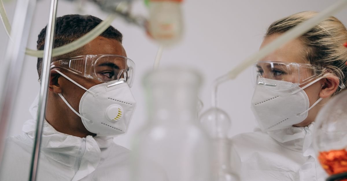Dois profissionais com máscara, óculos de proteção e traje branco trabalham em laboratório
