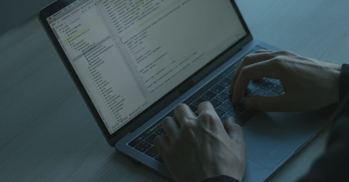 Mãos digitando em teclado de laptop com tela de código em ambiente com pouca luz