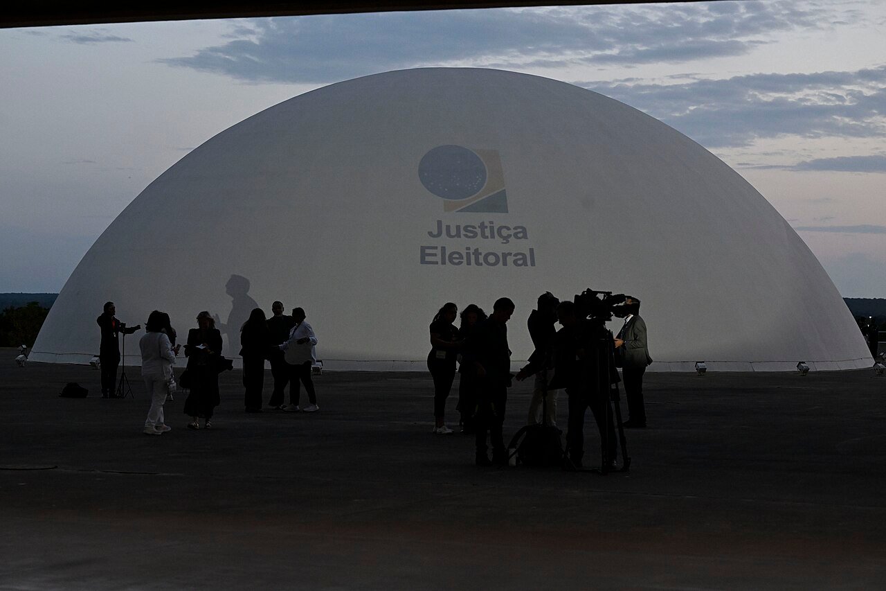Cúpula do Senado com a marca da Justiça Eleitoral projetada e equipes de imprensa em primeiro plano, na noite do primeiro turno de 2026