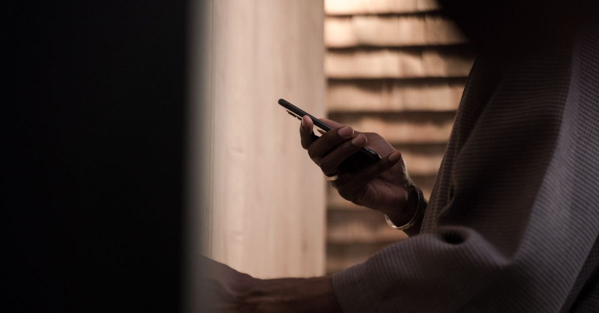 Pessoa segurando um celular em ambiente com pouca luz, com o rosto fora do enquadramento