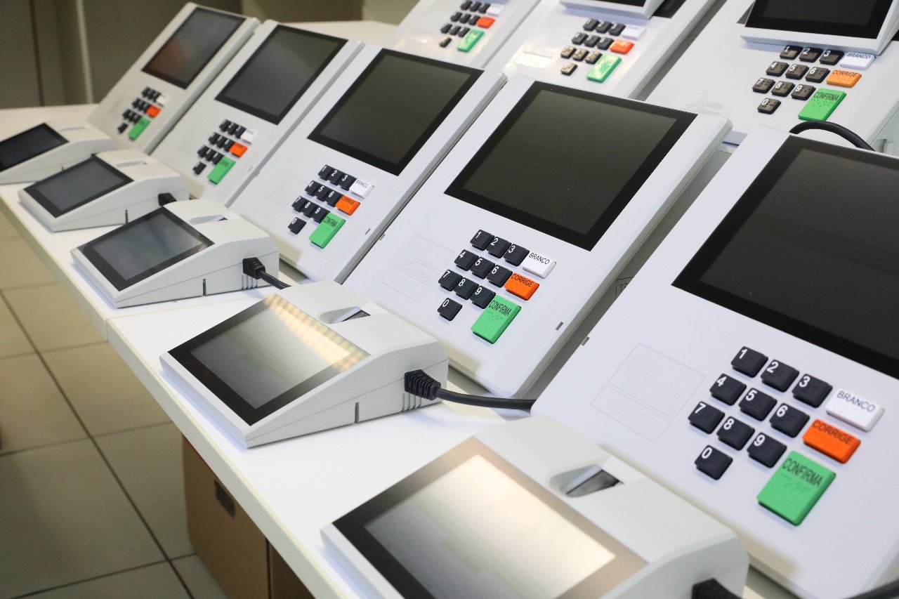 Urnas eletrônicas brasileiras enfileiradas sobre bancada, com teclado numérico e teclas branco, corrige e confirma