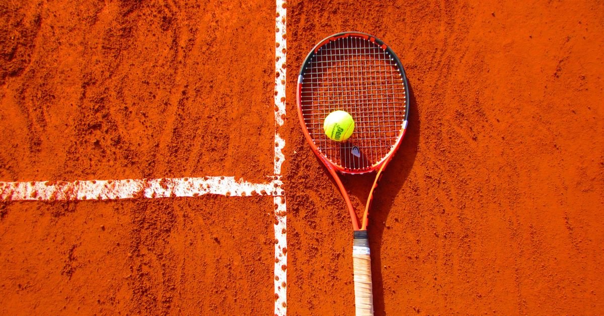 Raquete de tênis e bola amarela sobre quadra de saibro com linha branca